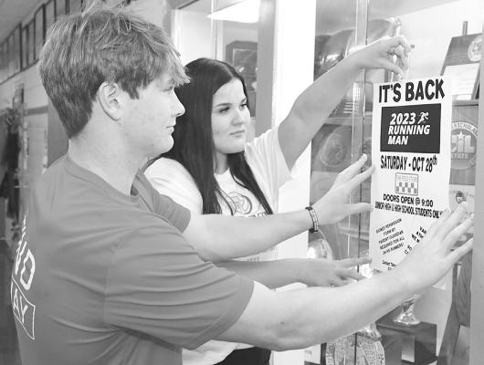Tyler Chapman (l) helps Cambree Clegg (r) tape up a poster promoting the 2023 Running Man. The Feed Store will host the popular event this Saturday night. Donnie Lucas / Albany News